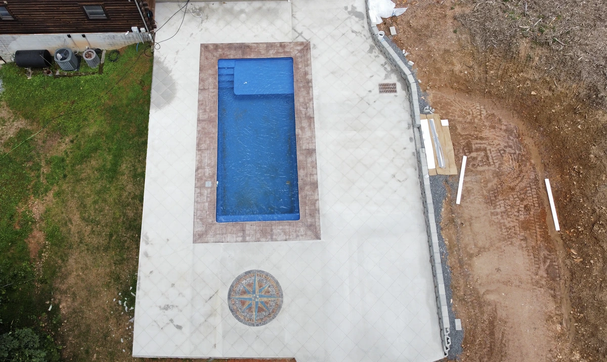 Aerial view of a rectangular pool ringed by stamped concrete coping and a wide scored pool deck with a compass rose medallion &mdash; The Concord Concrete Company