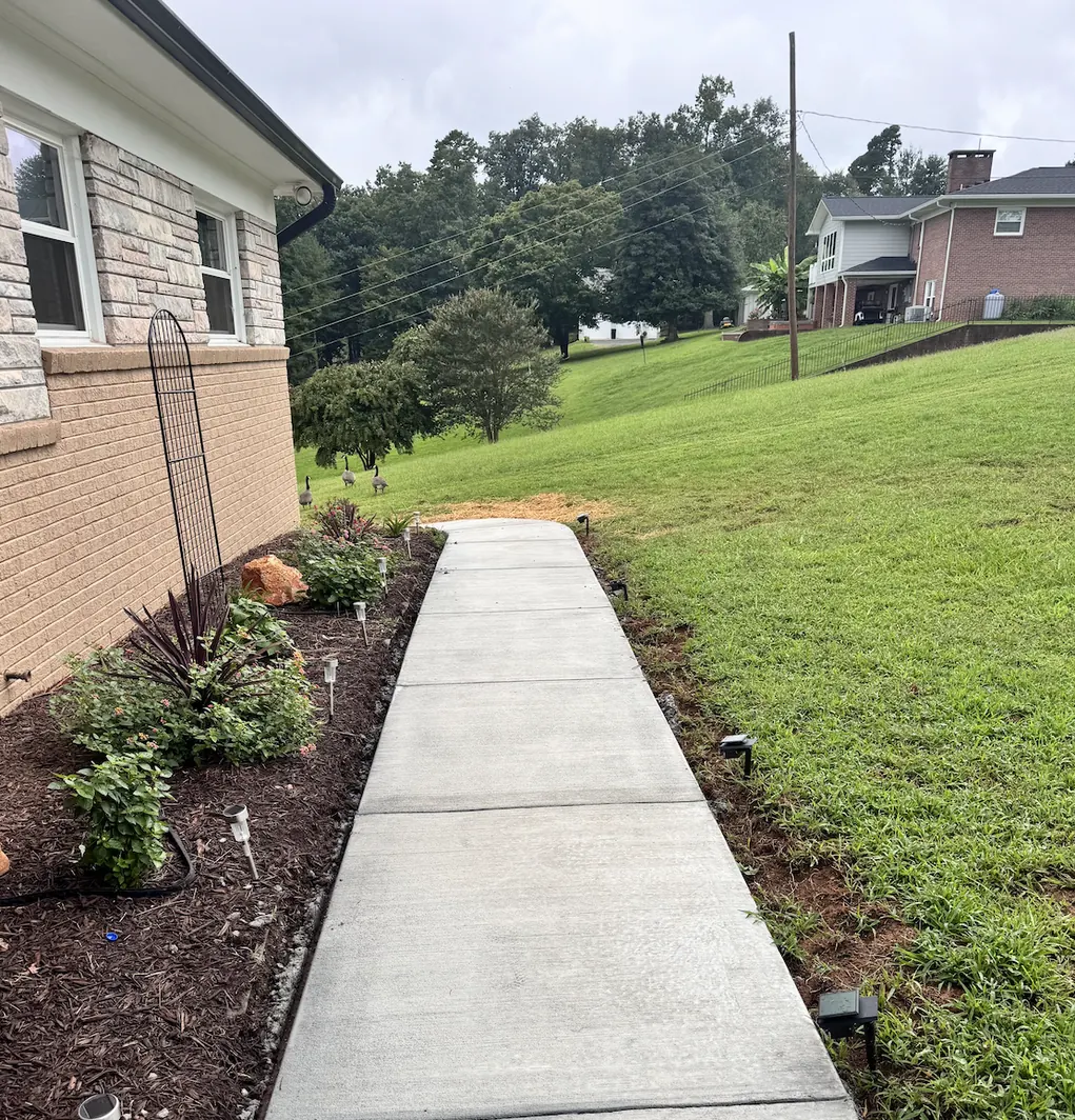 Straight concrete walkway with evenly spaced control joints beside a stone-front home, bordered by a planted bed and path lights &mdash; The Concord Concrete Company