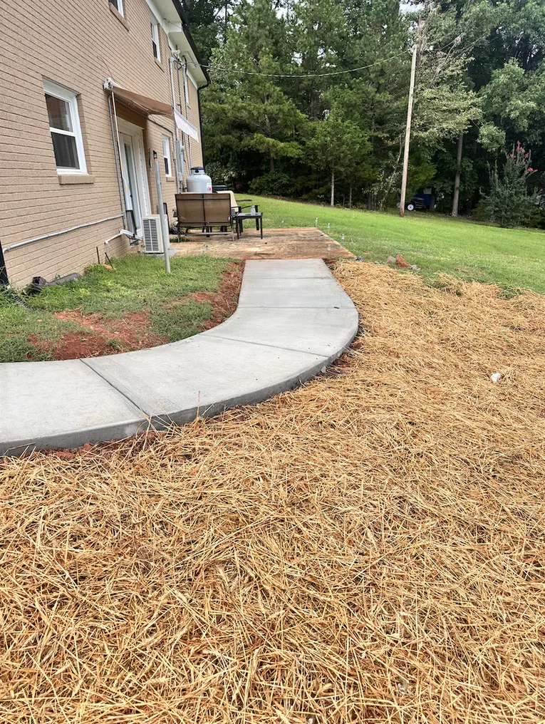Curved concrete walkway with tooled control joints running alongside a brick home to a rear patio, edged with fresh pine straw &mdash; The Concord Concrete Company