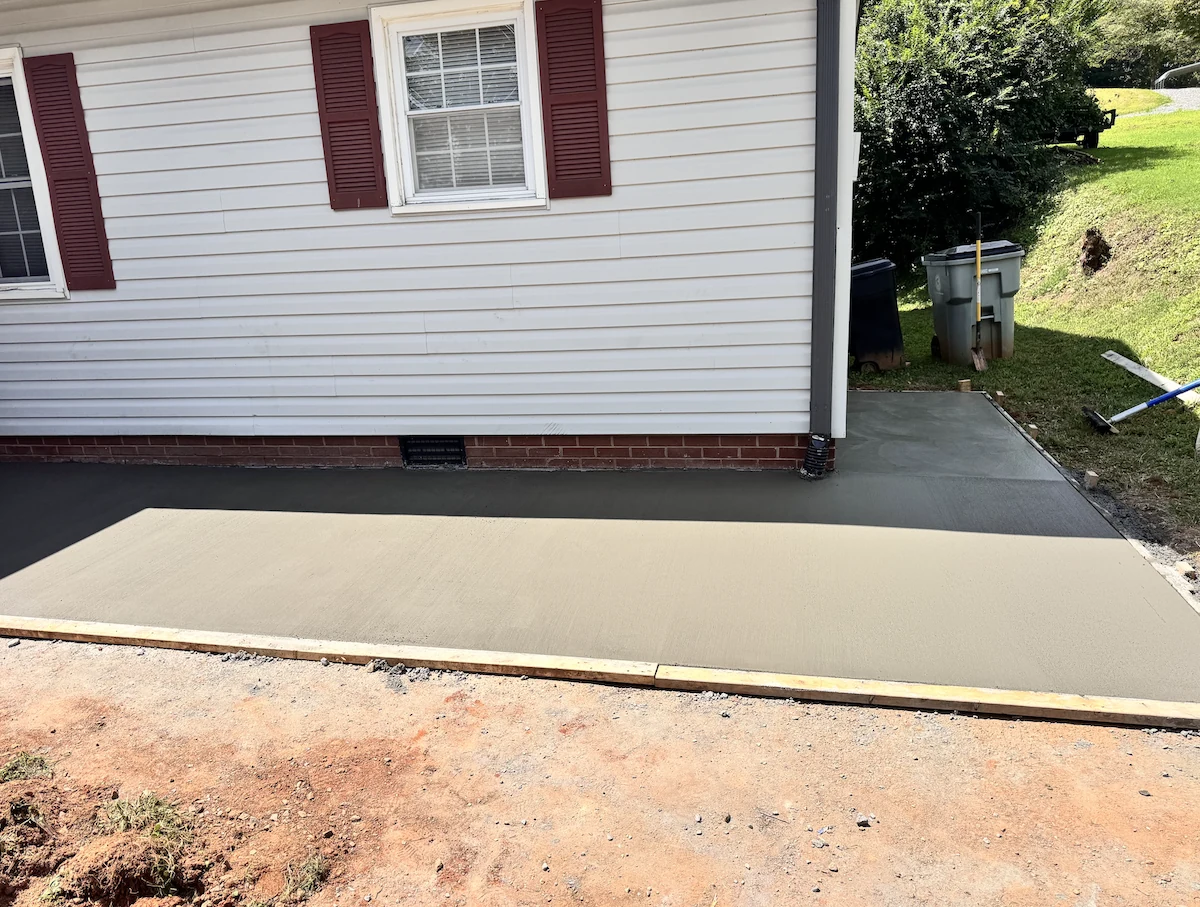 New concrete parking pad curing inside timber edge forms alongside a brick-skirted home in the NC Foothills &mdash; The Concord Concrete Company
