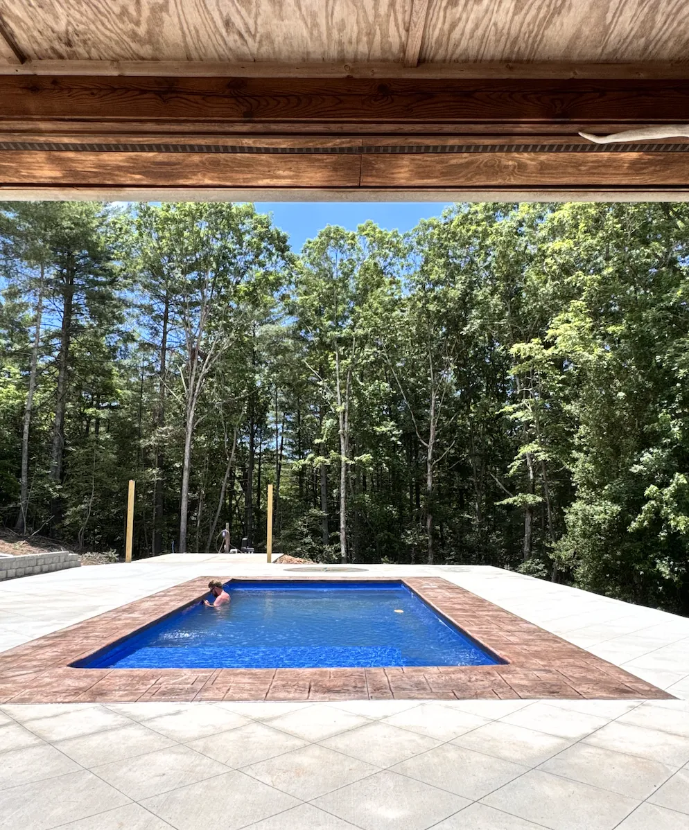 View from a covered porch across a wide stamped concrete pool deck to a rectangular pool with rust-toned stamped coping, a swimmer in the water and mature woods beyond &mdash; The Concord Concrete Company
